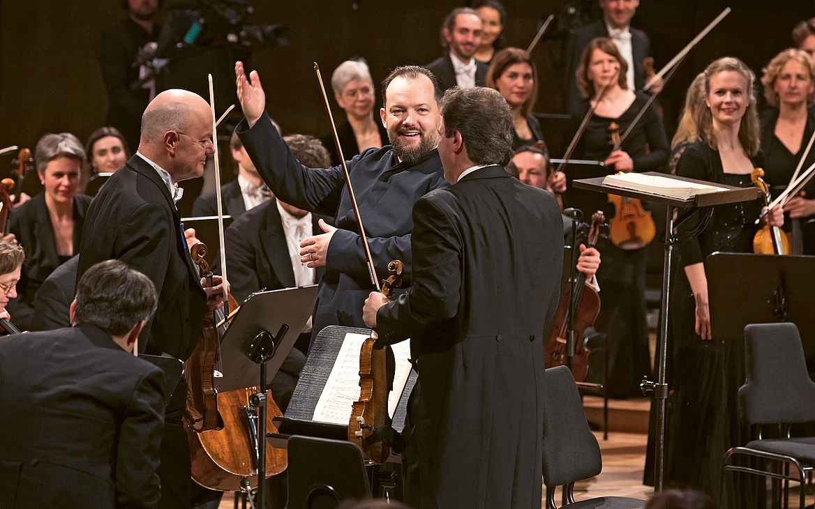 Das Foto zeigt Andris Nelsons, der in den Reihen der ersten Geigen des Gewandhausorchesters steht und die Musikerinnen und Musiker für den Schlussapplaus zum Aufstehen bittet. 