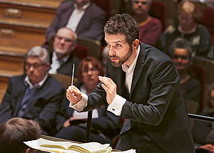 Das Foto zeigt den Dirigenten Omer Meir-Wellber von oben-vorne, der das Gewandhausorchester dirigiert. Hinter dem Dirigenten sind einige  Menschen im Publikum zu erkennen.