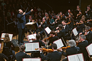 Das Foto zeigt den Dirigenten Andris Nelsons. Die Aufnahme ist über die bratschen hinweg geschossen, sodass diese im Bildvordergrund von hinten zu sehen sind. Der Dirigent dirigiert das Gewandhausorchester mit auf KOpfhöhe erhobenen nach vorne ausgestreckten Armaen. HInter ohm ist Publikum zu sehen, rechts von ihm und vor ihm erste Geigen und Celli.