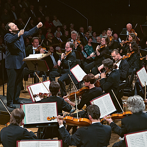 Das Foto zeigt den Dirigenten Andris Nelsons. Die Aufnahme ist über die bratschen hinweg geschossen, sodass diese im Bildvordergrund von hinten zu sehen sind. Der Dirigent dirigiert das Gewandhausorchester mit auf KOpfhöhe erhobenen nach vorne ausgestreckten Armaen. HInter ohm ist Publikum zu sehen, rechts von ihm und vor ihm erste Geigen und Celli.