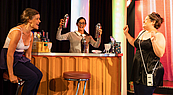 Auf dem Bild ist ein Bartresen abgebildet, hinter dem eine Dame mit zwei Getränkemixern in der Hand steht und in die Kamera lächelt, sowie zwei lachenden Damen vor dem Tresen.