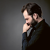 Das Bild zeigt den Dirigenten Petr Popelka im Profil bis zur Hüfte vor einer dunkelgrauen Wand. Er hat die Arme über dem Bauch verschränkt und stützt den nach unten geneigten Kopf dabei in die linke Hand.