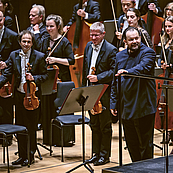 Das Foto zeigt dern Dirigenten Andris Nelsons, einige Musikerinnen und Musiker der ersten Geiden und der Celli. Sie stehen zum Publikum gewandt im Gewandhaus auf der Bühne und nehmen Applaus entgegen.