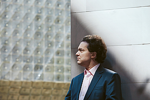Das Foto zeigt ein Brusportrait des Pianisten Evgeny Kissin. Er steht ein wenig nach rechts aus der MItte des Bildes gerückt, trägt ein weißes Hemd, einen mittelblauen Blazer und schaut zur linken Seite. Hinter ihm erheben sich zwei unterschiedlich strukturierte Weiße Wände, die sich in der MItte treffen.