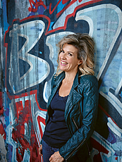 Das Foto zeigt die Geigerin Anne-Sophie Mutter, die lässig an einer Graffiti besprühte Wand gelehnt steht. Die Künstlerin dreht den Kopf zum Betrachter und lacht. Sie trägt eine blaue Bolero-Jacke und ein dunkelblaues Top.