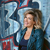 Das Foto zeigt die Geigerin Anne-Sophie Mutter, die lässig an einer Graffiti besprühte Wand gelehnt steht. Die Künstlerin dreht den Kopf zum Betrachter und lacht. Sie trägt eine blaue Bolero-Jacke und ein dunkelblaues Top.