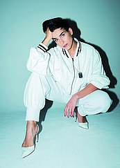 Das Foto zeigt die Sängerin Asmik Grigorian in einem weißen ANzug, in der Hocke mit nach vorne gestrecktem Bein, die KOpf auf eine Hand gestützt vor einer hellgrünen Wand.