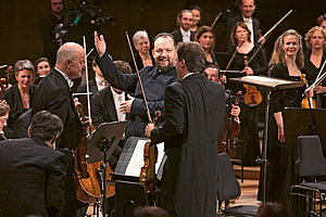 Das Foto zeigt Andris Nelsons, der in den Reihen der ersten Geigen des Gewandhausorchesters steht und die Musikerinnen und Musiker für den Schlussapplaus zum Aufstehen bittet. 
