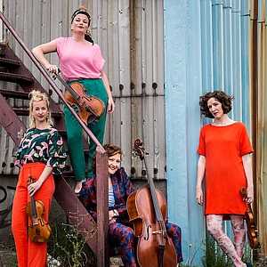Das Foto zeigt die vier Musikerinnen des Ragazze Quartetts vor einer INdustriefassade aus blauem Wellblech, sie tragen farbenfohe KLeidung in rot, grün, violoett und rosa