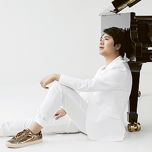 Das Foto zeigt den Pianisten Lang Lang in einem weißen raum, wie er in einem weißen Anzug am vorderen bein eines Flügels gelehnt auf dem Boden sitzt, ein Bein von sich gestreckt, weines angewinkelt, auf dem sein UNterarm ruht.