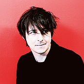 Das Foto zeigt ein Portrait des Pianisten Michael Wollny mit wirr in die STirn fallenden Haarn vor einer roten Wand. Er trägt eine schwarze Jacke und ein schwarzes T-Shirt.