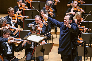 Das Foto zeigt den Dirigenten Andris Nelsons, der das Gewandhausorchester im Gewandhaus dirigiert. Er steht am rechten Bildrand, nach links schauend, die Arme weit nach vorne, halb seitlich ausgestreckt. HInter ihm sind Musiker der zweiten  Geigen zu sehen, vor ihm Musiker der Bratschen