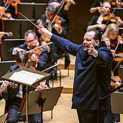 Das Foto zeigt den Dirigenten Andris Nelsons, der das Gewandhausorchester im Gewandhaus dirigiert. Er steht am rechten Bildrand, nach links schauend, die Arme weit nach vorne, halb seitlich ausgestreckt. HInter ihm sind Musiker der zweiten Geigen zu sehen, vor ihm Musiker der Bratschen