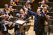 Das Foto zeigt den Dirigenten Andris Nelsons, der das Gewandhausorchester im Gewandhaus dirigiert. Er steht am rechten Bildrand, nach links schauend, die Arme weit nach vorne, halb seitlich ausgestreckt. HInter ihm sind Musiker der zweiten Geigen zu sehen, vor ihm Musiker der Bratschen