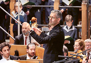 Das Foto zeigt ein Halbkörperfoto des Thomaskantors Andreas Reize im Profil in Nahaufnahme beim Dirigieren in der Thomaskirche. Im Hintergrund sind einige Sänger des Thomanaerchores zu sehen und einge Musiker des gewandhausorchesters.