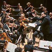 Das Foto zeigt den Dirigenten Andris Nelsons von der Seite, der das Gewandhausorchester im Leipziger Gewandhaus dirigiert. Vom Orchester sind die zweiten Geigen, die Bratschen und die ersten Celli zu sehen.