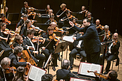 Das Foto zeigt den Dirigenten Andris Nelsons von der Seite, der das Gewandhausorchester im Leipziger Gewandhaus dirigiert. Vom Orchester sind die zweiten Geigen, die Bratschen und die ersten Celli zu sehen.