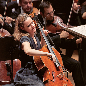 Das Foto zeigt eine Nahaufnahme der Cellistin Sol Gabetta im Gewandhaus, die leidenschaftlich ihr Instrument spielt. Im Hintergrund sind wneige Musiker des Gewandhausorchesters zu sehen.