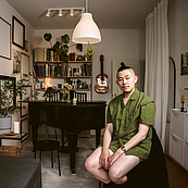 Das Foto zeigt den Taiwanesischen Dramaturgen Ching-Tien Lin in seinem Zuhause. Im Hintergrund steht ein Flügel mit dem Endstück zum Betrachter zeigend. Lin trägt ein grünes Hemd und kurze Hosen.