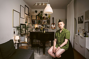 Das Foto zeigt den Taiwanesischen Dramaturgen Ching-Tien Lin in seinem Zuhause. Im Hintergrund steht ein Flügel mit dem Endstück zum Betrachter zeigend. Lin trägt ein grünes Hemd und kurze Hosen.