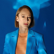 Das Foto zeigt die Astrophysikerin Sibylle Anderl in einem Blauen Jackett vor einer blauen Wand stehend. Eine weiße LIchtreflexion wie Wolkenstreifen bedeckt ihren Oberkörper und die Wand